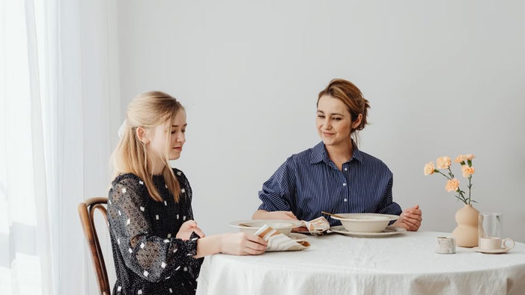 A mother and daughter sit at a table with flowers, enjoying dinner in a cozy indoor setting.
